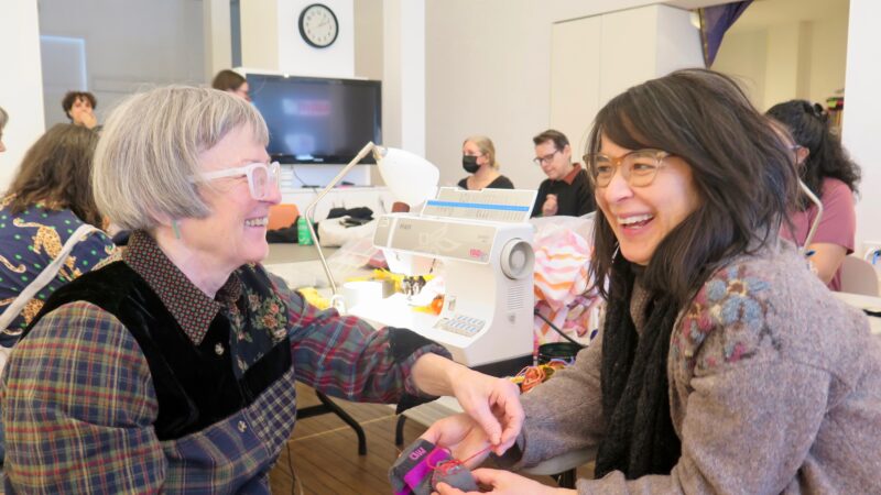 Two people sitting in front of a sewing machine smiling as they talk.