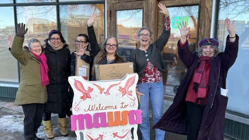 A group of six women stand on the sidewalk outside MAWA's old Main St. location, waving.
