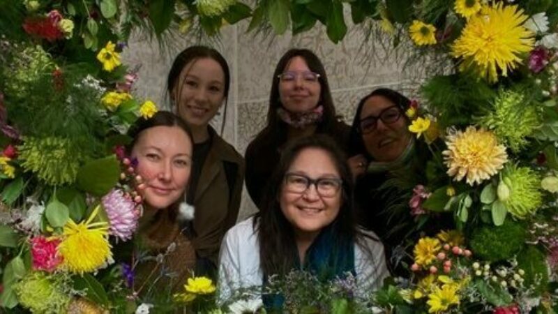 The members of the Mîcêt Tipiskâwi-pîsimak Collective, Frances Cooper, Annie Courchene , Tess Ray Houston, Lita Fontaine and Kristy Janvier, look through the center of a very large lush flower wreath.