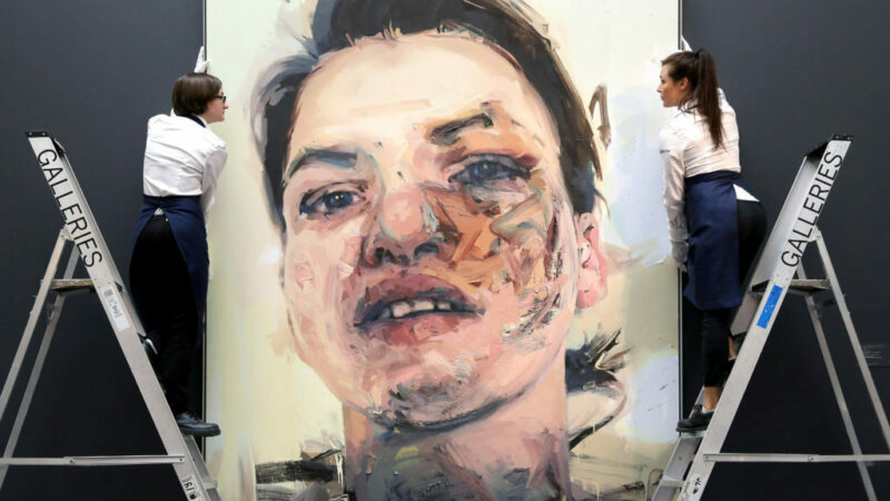 Two people on ladders adjust a large painting by Jenny Saville. The painting is a portrait rendered in a loose style.