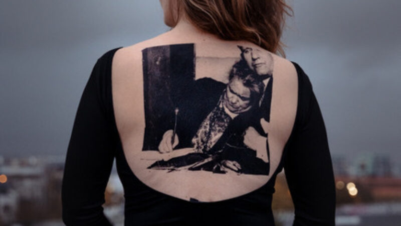 A woman stands with her back to the camera wearing a black shirt that leaves her back bare. An image is imprinted on her skin.
