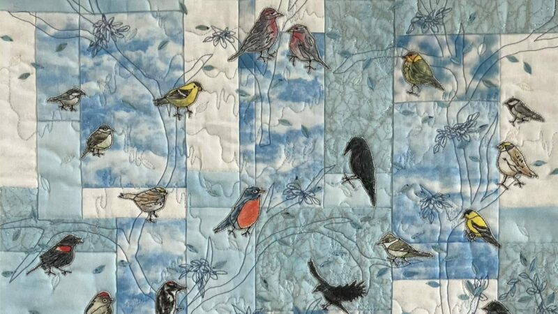Textile art piece by Cathie Ugrin with squares of blue and white fabric, embroidered branches and appliqued birds.