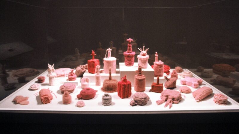 An installation of numerous knitted reproductions of various anti personnel mines in shades of pink on a white platform in a darkened gallery.