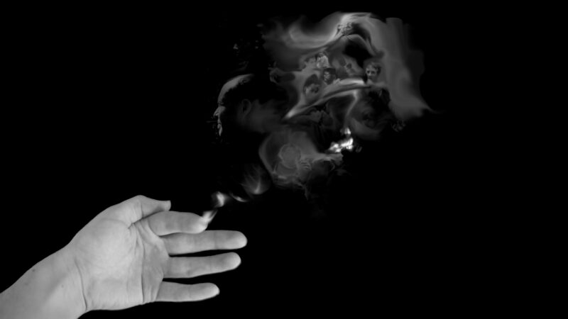 Black and white photo of a hand with a burning fingertip. In the smoke from the fingertip are many ghostly faces.