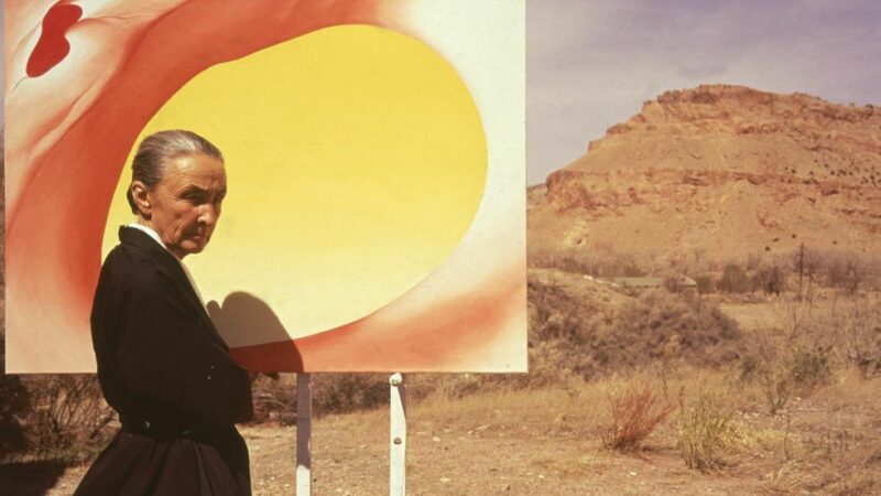 Georgia O'Keefe stands in front of a canvas on an easel in a desert landscape.