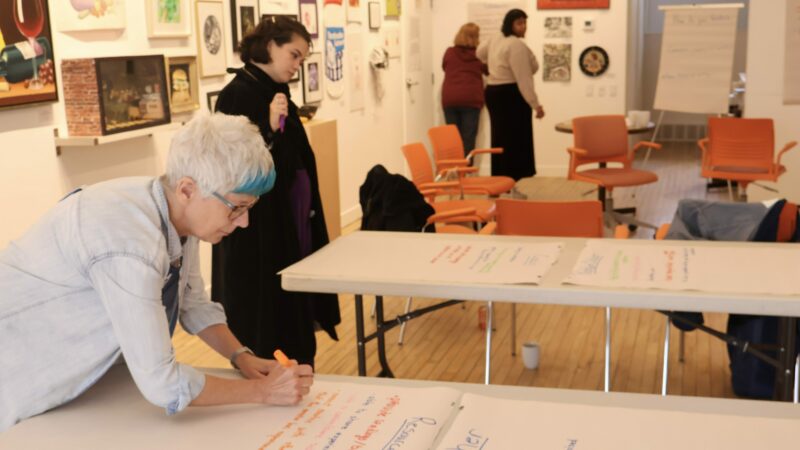 A person leans over to write on a large piece of paper laid out on a table. Other workshop participants are seen in the background.