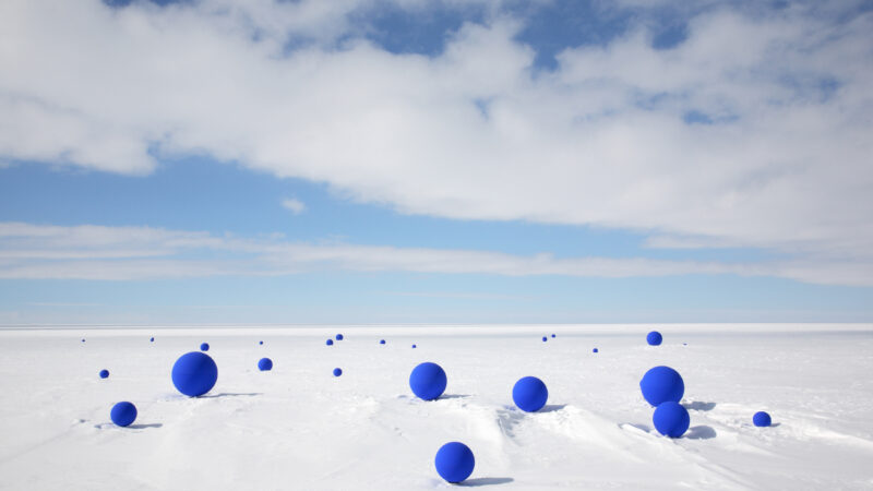Blue sphere of various sizes sit scattered across a flat snowscape under a blue sky.