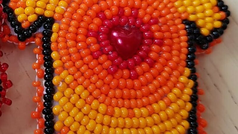 A beaded pin in the shape of a t-shirt. In the center of the shirt is a heart shaped bead and the rest of the orange and yellow beads radiate out from the heart.