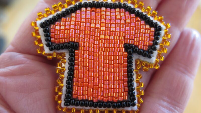 A beaded pin of an orange shirt outlined in black beads with a gold trim sits in the palm of a person's hand.