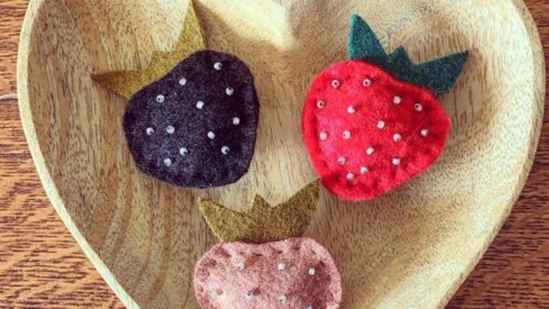 Three felt strawberries in dark purple, red and light pink with green tops and beads as seeds sit in a wooden heart shaped bowl.