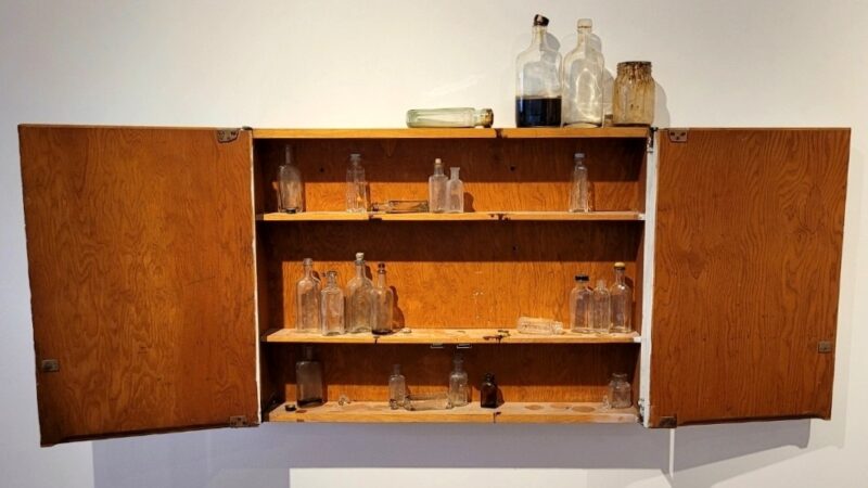 An open wooden medicine cabinet on a white wall with small glass medicine or liquor bottles inside.