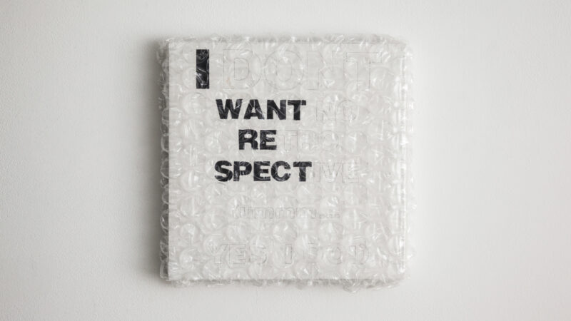 A square white canvas wrapped in bubble wrap hangs on a white wall. Black block letters say I WANT RE SPECT other letter outlined in light pencil are present but not legible.