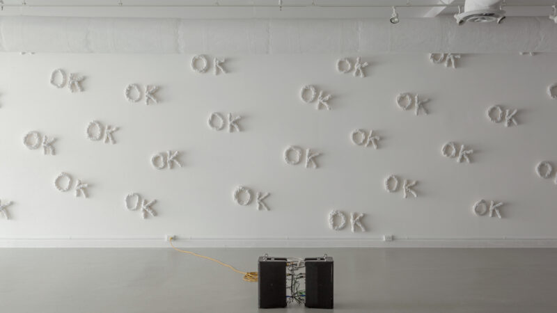 A photo in a gallery of an installation artwork by Tara Lynn MacDougall. The word OK in 3-D sculpted out of white material is repeated over and over on a white wall. On the floor are two speakers.