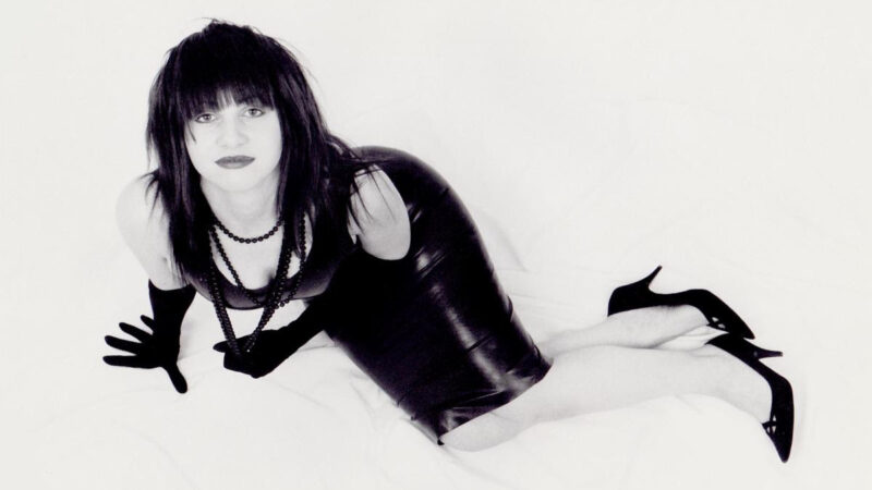 Black and white photo of Lydia Lunch wearing a black dress, high heels and gloves.  She is lying on a white background looking up at the camera.