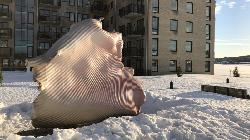 A large sculpture made of a sheet of metal formed into a wavy somewhat organic shape sits outside in the snow.
