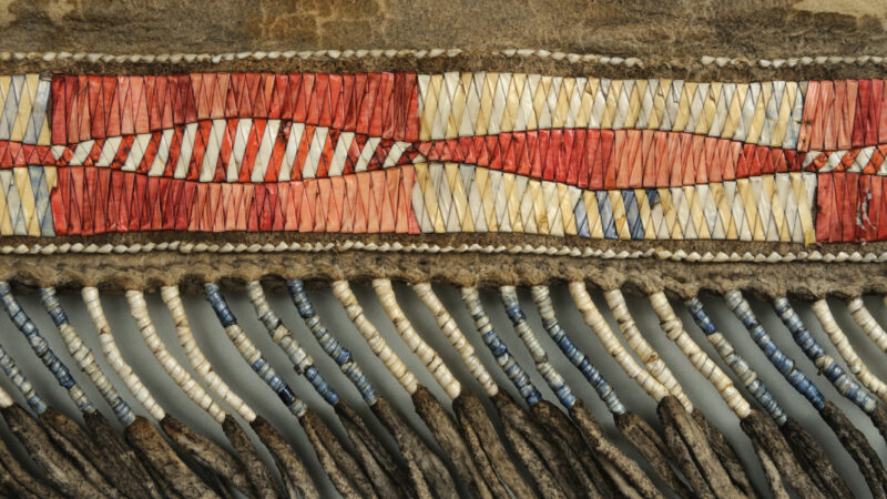 Detail of a wrapped fringe from a man's legging made using quillwork. The quills are coloured red and white and are woven into a geometric pattern.