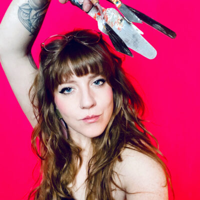 Photo of Laura Lewis in front of a pink background looking at the camera while holding palette knives over her head. Lewis has fair skin and long wavy blond hair with bangs.