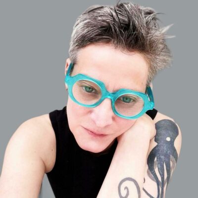 Photo of Chantal Nadeau with her head tilted forward and to the right as if she if looking at a screen in front of her. She has light skin and short hair. She wears glasses with thick aqua frames, a black tank top and has a tattoo of an octopus on her right arm.
