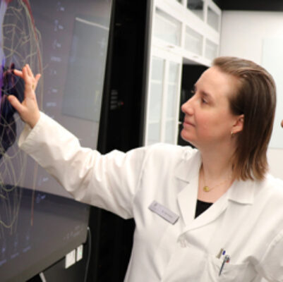 Photo of Dr. Sari Hannila in a white lab coat reaching up to touch a large screen showing brain imaging.