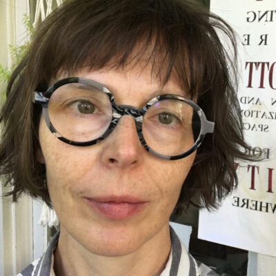 Photo of hannah_g. She has light skin and chin length brown hair with bangs. She is wearing glasses with black and grey frames and a striped white and grey button up shirt.