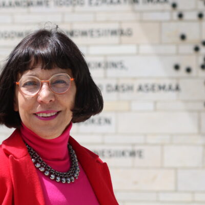Headshot of Serena Keshavjee. Keshavjee is looking a the camera smiling. Her chin length black hair blows slightly in the wind. She has light skin and wears glasses, a red blazer and pink turtleneck.