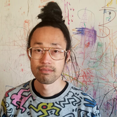 Photo of Takashi Iwasaki standing in front of a wall covered with paint splatters. He is wearing a shirt with a Keith Harring design.
